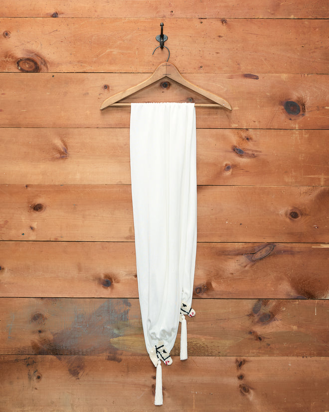 Sequin Pony Scarf in Cream with silken tassels hangs on a wooden hanger against a rustic wooden wall.
