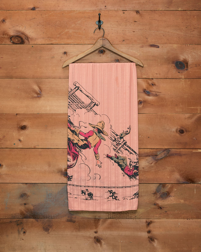 The Bronco Buster Scarf, a pink towel featuring cartoon cowboys reminiscent of a cowboy scarf, hangs on a wooden wall on a hanger.