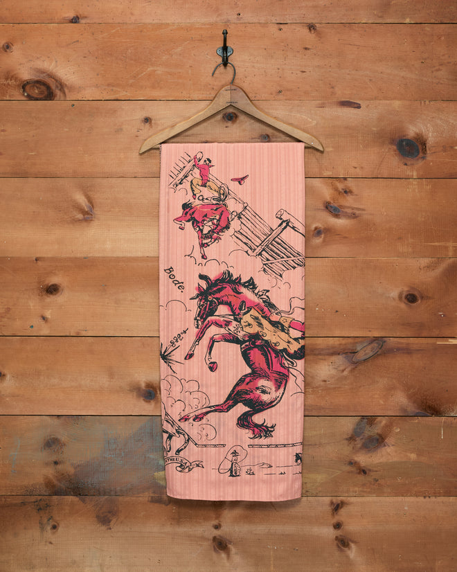 Bronco Buster Scarf featuring lively illustrations of rodeo scenes and bucking horses, reminiscent of the vibrant rodeos in Cheyenne, Wyoming, hanging on a wooden wall.