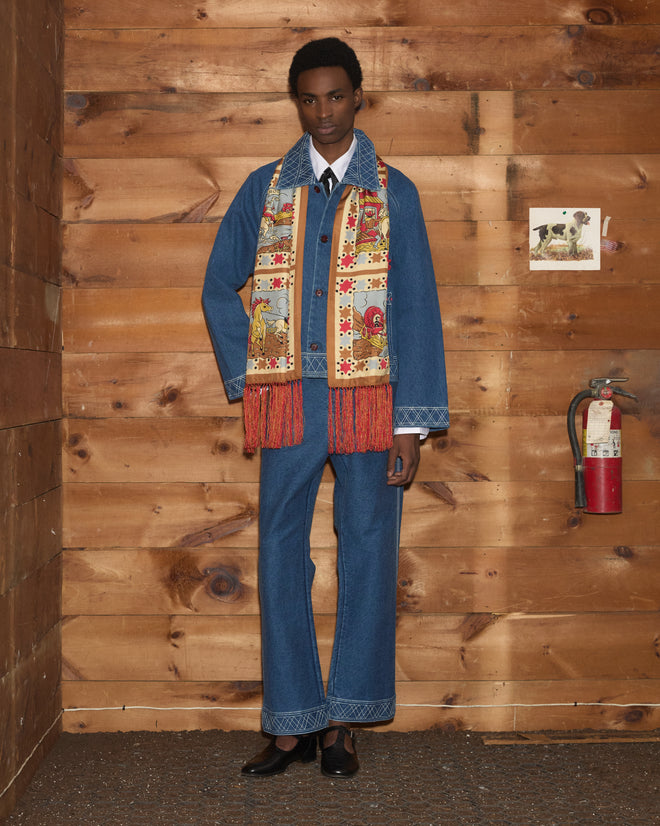 Wearing the Cowboy Comic Scarf with a denim suit, the individual stands indoors by a wooden wall and fire extinguisher, channeling the playful charm of a classic cowboy comic.