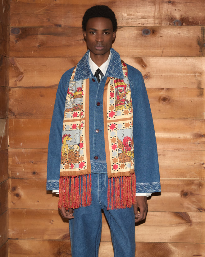 Wearing the Cowboy Comic Scarf—a colorful silk accessory with a red fringe—a person in a denim jacket and jeans stands against a wooden wall, evoking the style of a Western comic scene.