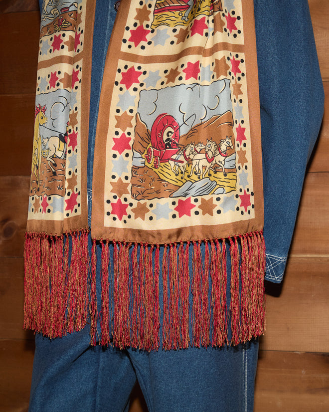 A person in a blue denim outfit wears the Cowboy Comic Scarf—featuring cowboy-themed comic prints and red fringe—while standing by wood paneling.