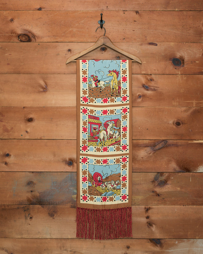 Cowboy Comic Scarf featuring three cartoon cowboy comic scenes, star borders, and a red fringe along the bottom, displayed on a wooden background.