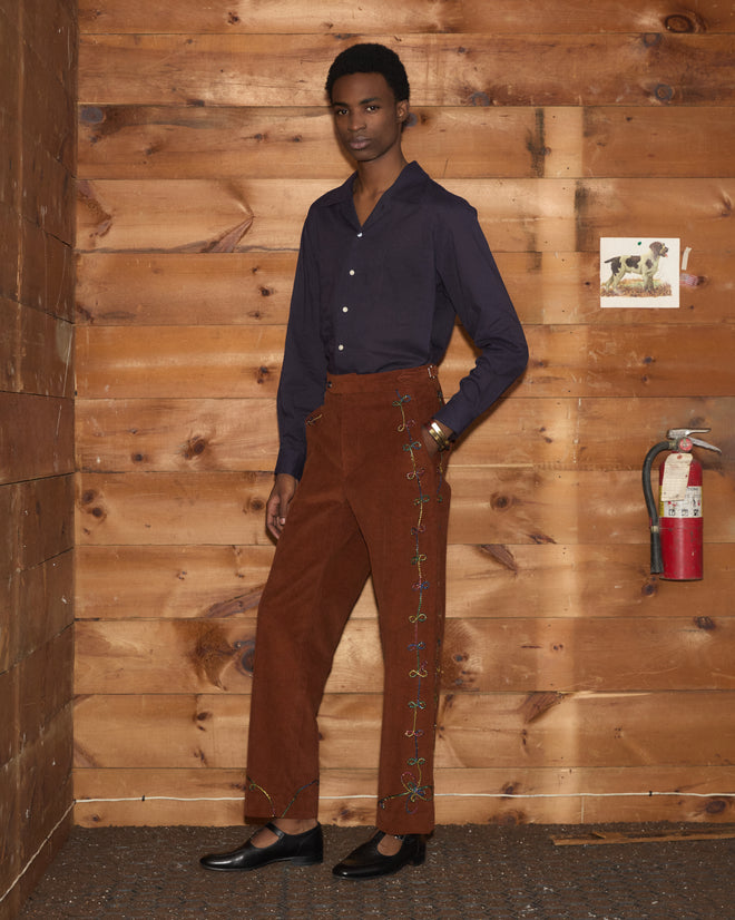 A person wearing a navy shirt and Cactus Couching Trousers stands in a wooden room near a fire extinguisher and small wall art.