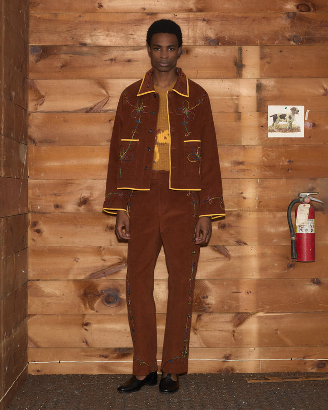 Person in the Cactus Couching Jacket with brown Western chain-stitch embroidery and black boots stands against a wooden wall with a fire extinguisher.
