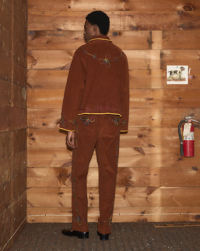 A person wearing the Cactus Couching Jacket stands facing a wooden wall, next to a fire extinguisher and a small picture. The embroidered outerwear showcases subtle Western chain-stitching that adds distinct character to the ensemble.