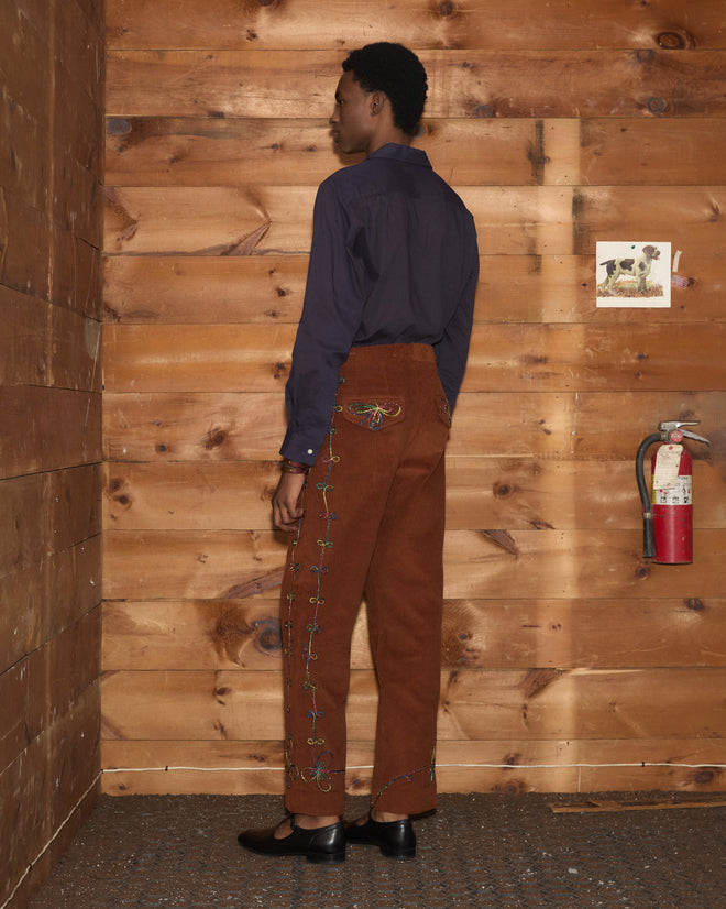 Person in Cactus Couching Trousers featuring rainbow embroidery and a navy shirt stands by a wooden wall with art and a fire extinguisher.