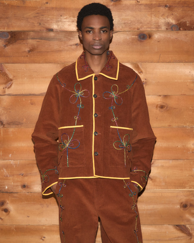 A person wearing the Cactus Couching Jacket, a brown embroidered corduroy piece, stands in front of a wooden wall with hands in pockets.