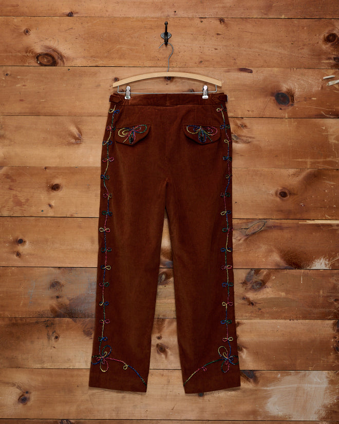 Cactus Couching Trousers—brown corduroy pants featuring vibrant rainbow embroidery and Western chain-stitching—hung on a hanger against a wood wall.