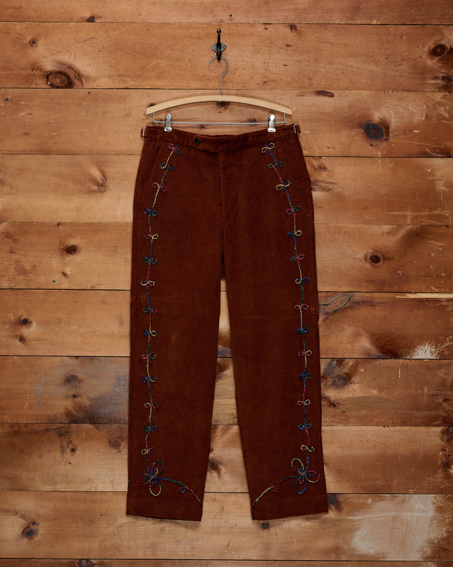 Cactus Couching Trousers in brown corduroy, featuring colorful rainbow embroidery and Western chain-stitching along the sides, displayed on a clear hanger against a wood wall.