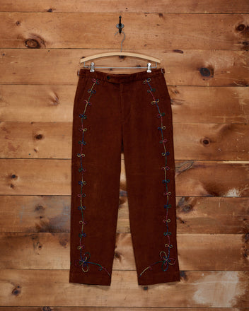 Cactus Couching Trousers in brown corduroy, featuring colorful rainbow embroidery and Western chain-stitching along the sides, displayed on a clear hanger against a wood wall.