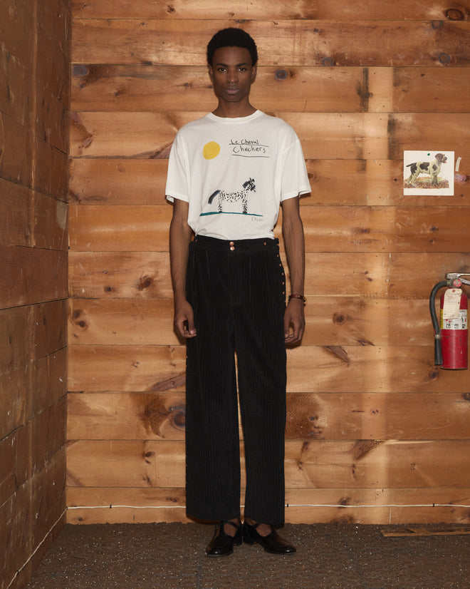 Person standing indoors against a wood wall, wearing the Cheval Tee made from 100% cotton, along with black pants and black shoes.