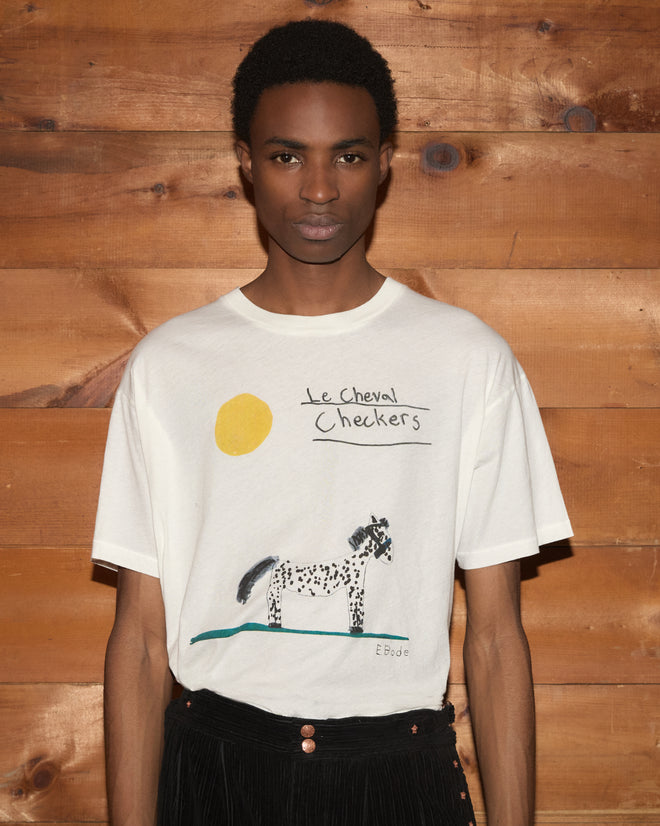 Wearing the Cheval Tee—a white 100% cotton T-shirt featuring a playful, childlike drawing of a spotted horse, sun, and the text “Le Cheval Checkers”—perfect for pony t-shirt lovers.