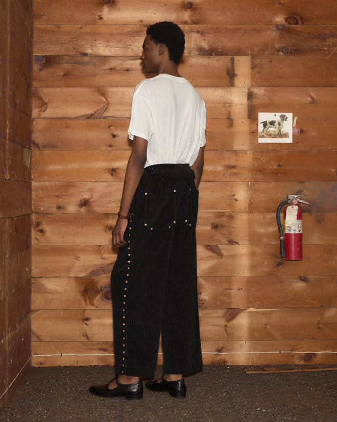 Person wearing Corduroy Copper Spur Trousers, a white t-shirt, and wide-leg black pants with floral side seams.