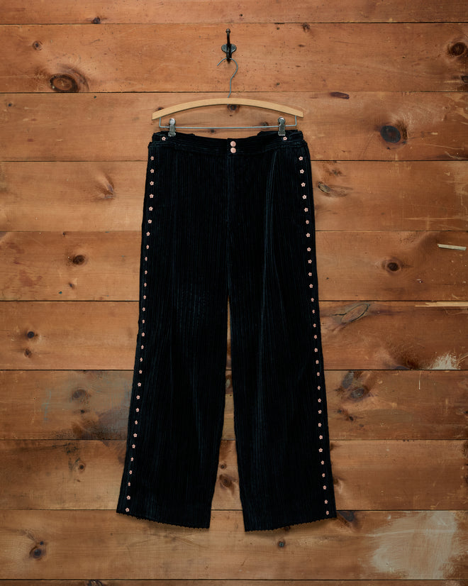 Corduroy Copper Spur Trousers: black straight-leg trousers with flower-shaped side buttons, on a wooden hanger.