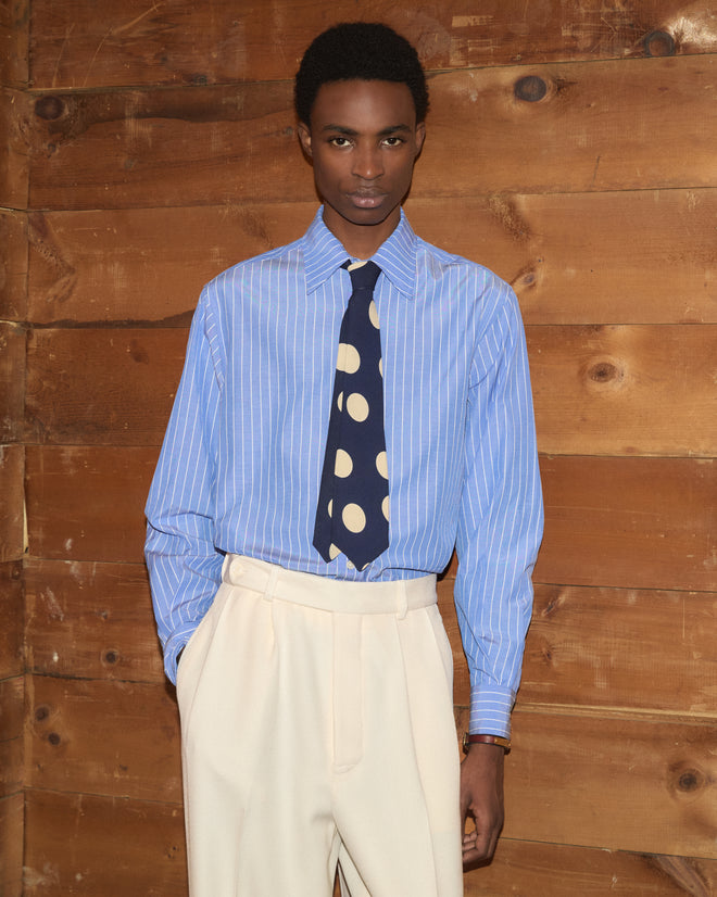 A model wears a Dot Crêpe Neck Tie in navy blue with cream dots over a blue and white striped shirt.