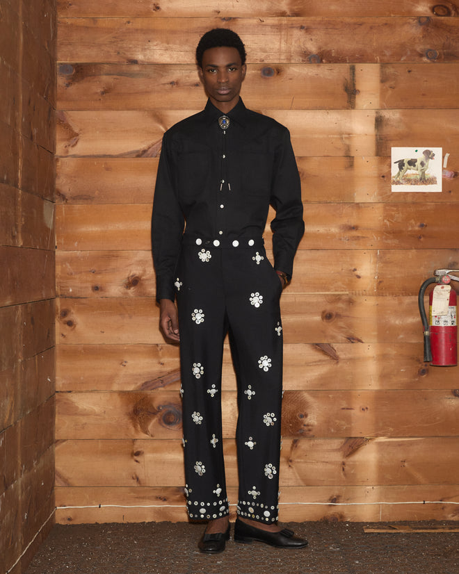 A person wears a black button-down, bolo tie, and Pearly Royal Side-Buckle Trousers with metallic floral studs.