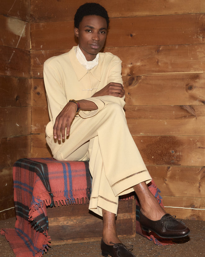A person in pale yellow Ladder Crêpe Pajama Pants and dark loafers, sitting on a wooden crate.