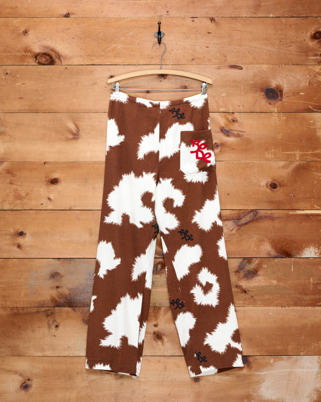 Flannel Cow Print Pajama Pants featuring a brown and white cow pattern with a white pocket, displayed hanging on a wooden wall.