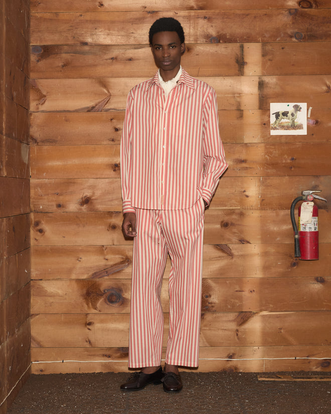 A person in a red and white striped pajama-style outfit stands by a wooden wall adorned with art and a fire extinguisher, their look completed by relaxed Rancho Pajama Pants made from 100% cotton.
