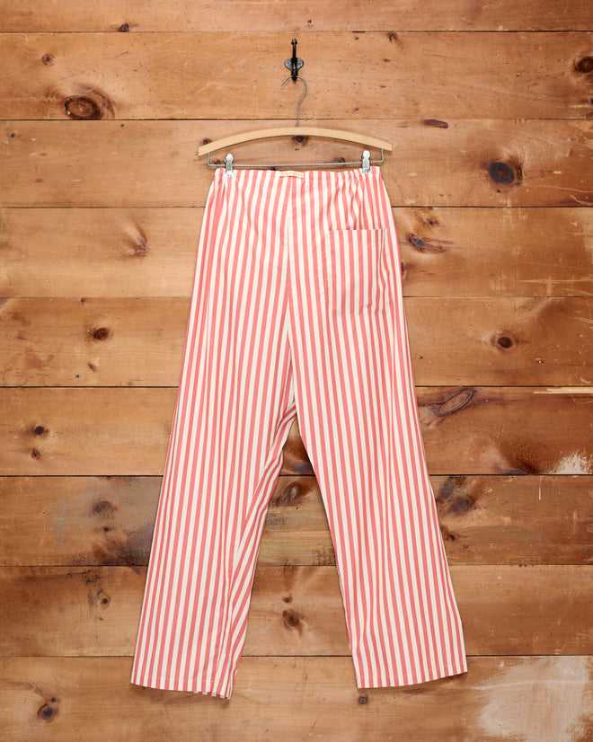 The Rancho Pajama Pants, featuring red and white vertical stripes and crafted from 100% cotton, hang on a wooden hanger against a rustic wooden wall background.