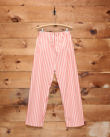 Rancho Pajama Pants, crafted from 100% cotton in a red and white striped design, hang on a wooden hanger against a rustic wooden wall.