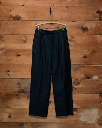 Mohair McNab Trousers: black wide-leg pleated trousers hanging on a wooden hanger against a wooden wall.