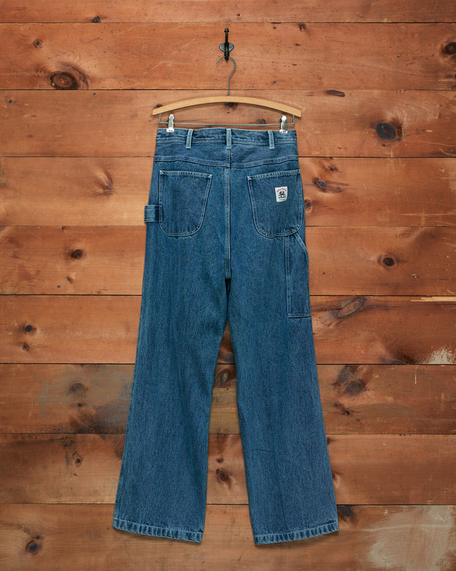 Washed Denim Knolly Brook Trousers hanging on a wooden hanger, set against a rustic wooden wall backdrop.