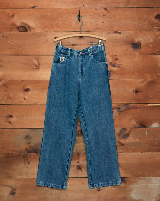 The Washed Denim Knolly Brook Trousers hang on a wooden hanger against a rustic wooden plank wall background.