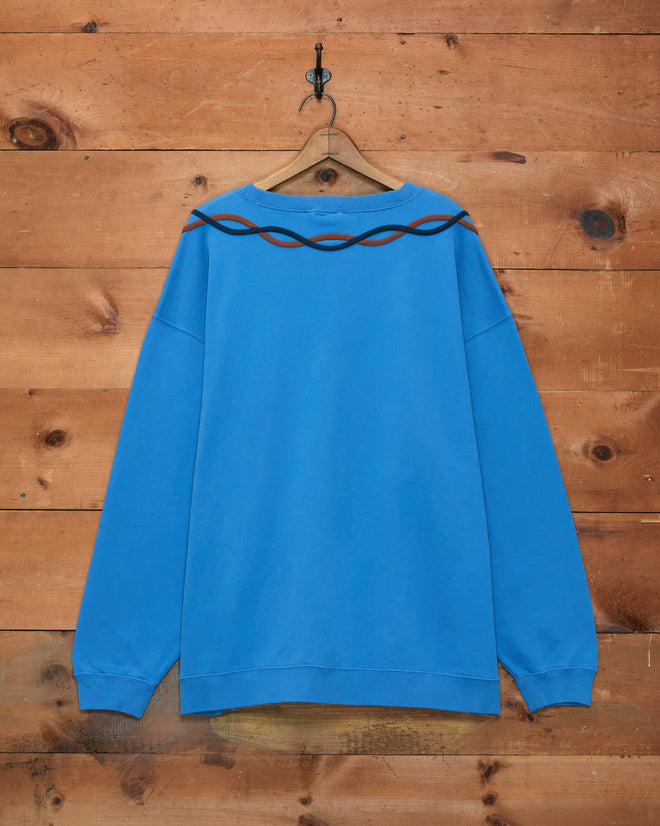 Sidewinder Pullover displayed on a wooden wall—a blue fleece sweatshirt featuring black and brown wavy line accents near the shoulders and collar. Inspired by 1960s sportswear, it is crafted from 100% cotton for a comfortable fit.
