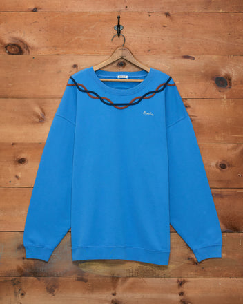 Sidewinder Pullover—blue fleece sweatshirt with brown wavy stripe detail inspired by 1960s sportswear, hanging on a wooden wall.