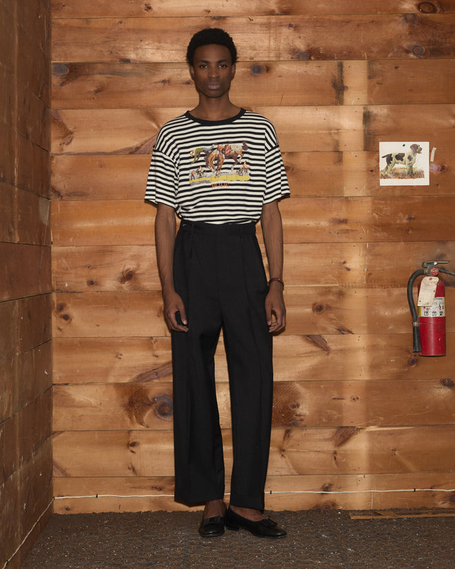 A person stands indoors, wearing the Rib Stripe Rodeo Tee and black pants, with a fire extinguisher mounted on the wooden wall behind them.