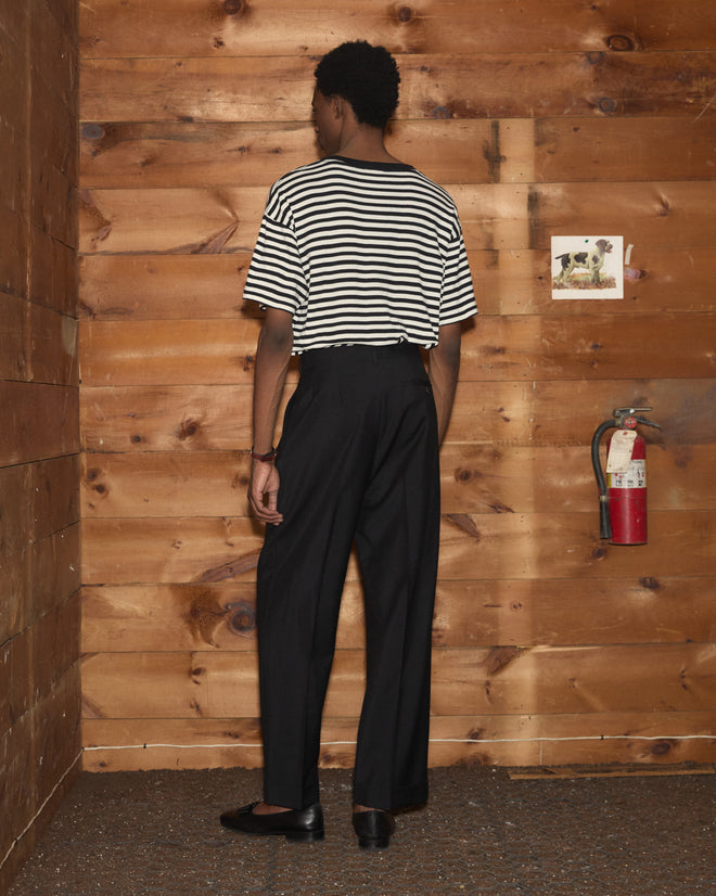 Wearing the Rib Stripe Rodeo Tee and black pants, a person stands facing a wooden wall with a fire extinguisher mounted on it.