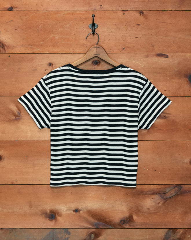 Rib Stripe Heart Tee featuring black and white stripes and short sleeves, displayed on a wooden hanger against a wooden wall.