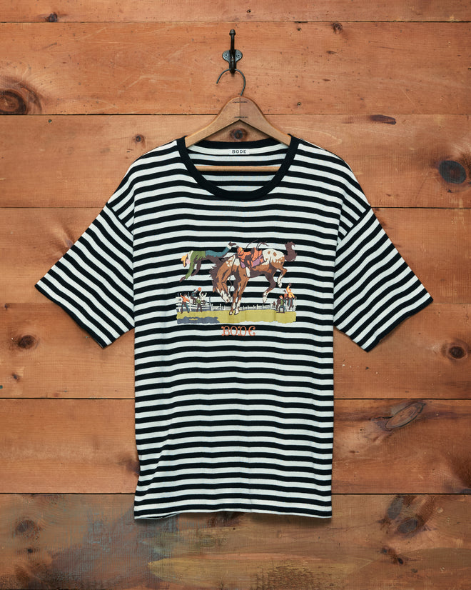 Rib Stripe Rodeo Tee, a black and white striped shirt with a horse riding graphic, crafted from 100% cotton with a subtle ribbed pattern, displayed hanging on a wooden wall.