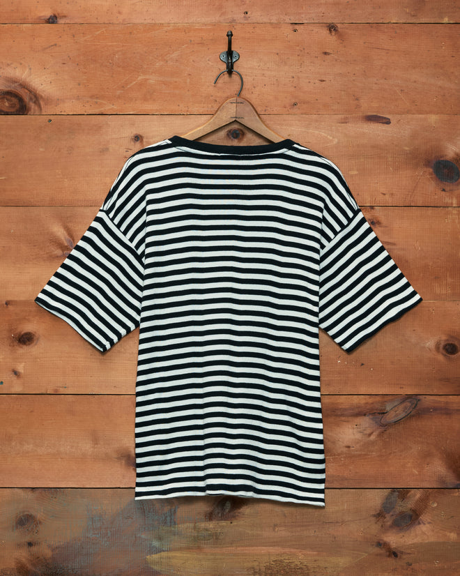 The Rib Stripe Rodeo Tee, crafted from 100% cotton with black and white stripes and short sleeves, is displayed hanging on a wooden wall.