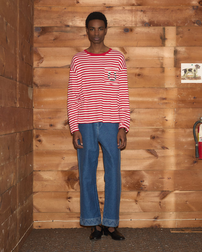 Wearing the Rib Stripe Horseshoe Long Sleeve Tee and blue jeans, a person stands against a wooden wall near a fire extinguisher.
