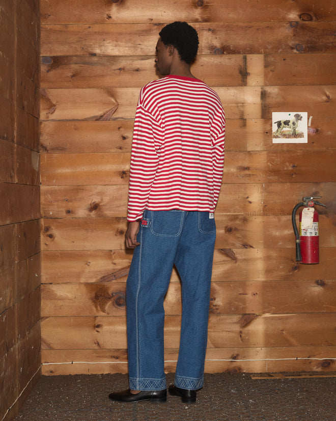 A person wearing the Rib Stripe Horseshoe Long Sleeve Tee and blue jeans stands facing a wooden wall with a fire extinguisher and a dog photo.