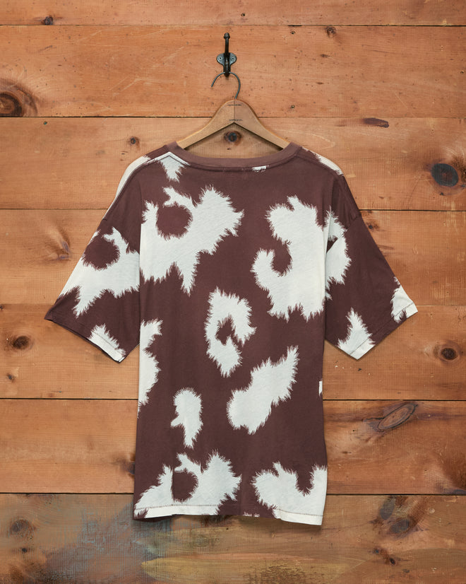 Featherweight Cow Print Tee, a brown and white tie-dye shirt with a cow print design, hanging on a wooden hanger against a wooden plank wall.