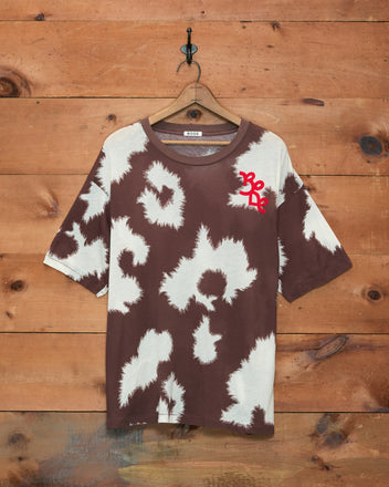 Featherweight Cow Print Tee featuring a brown and white tie-dye design reminiscent of cow print, with a red graphic on the left chest, displayed hanging on a wooden wall.
