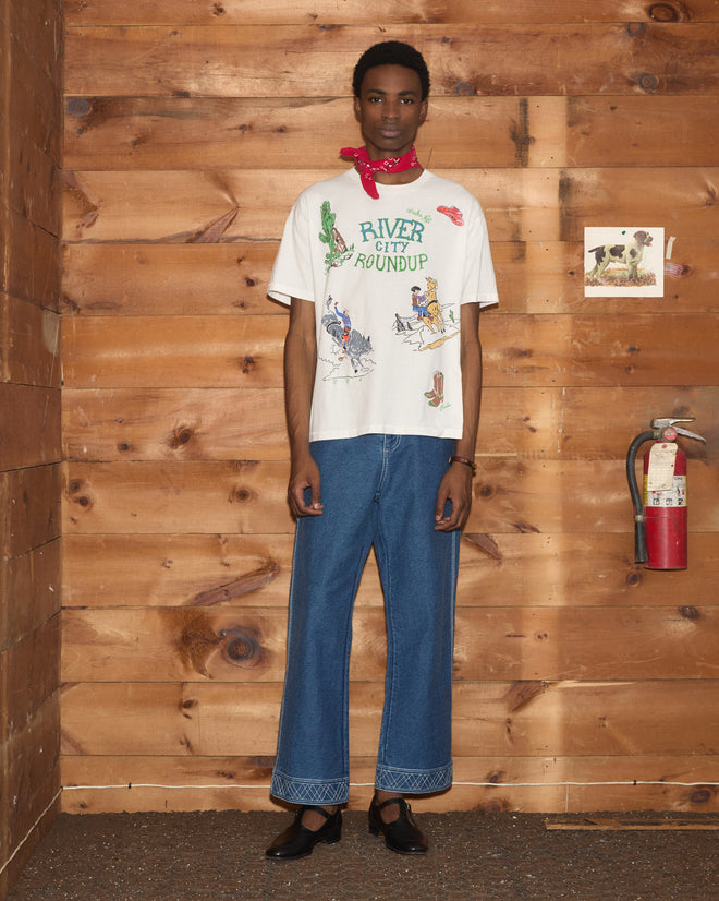 Wearing wide jeans, a River City Tee, a red scarf, and black shoes, this person stands by a wood wall with a fire extinguisher—ready for the River City Roundup or an Omaha rodeo.