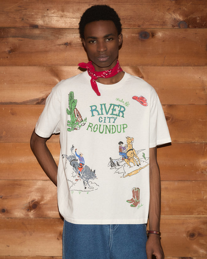 Wearing the River City Tee with cowboy illustrations, paired with a red bandana and blue jeans, this look channels the spirit of the Omaha rodeo against a wood wall backdrop.