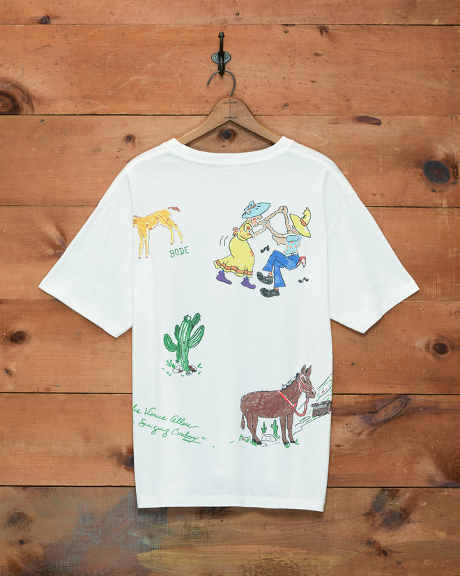River City Tee—a white T-shirt featuring cartoon cowboy scenes, cacti, and a donkey illustration, inspired by the spirit of the River City Roundup and Omaha rodeo, hanging on a wooden wall.