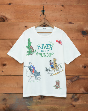 The River City Tee showcases rodeo-themed illustrations and “River City Roundup” lettering, hanging on a wooden wall—perfect for celebrating Omaha’s rodeo heritage.