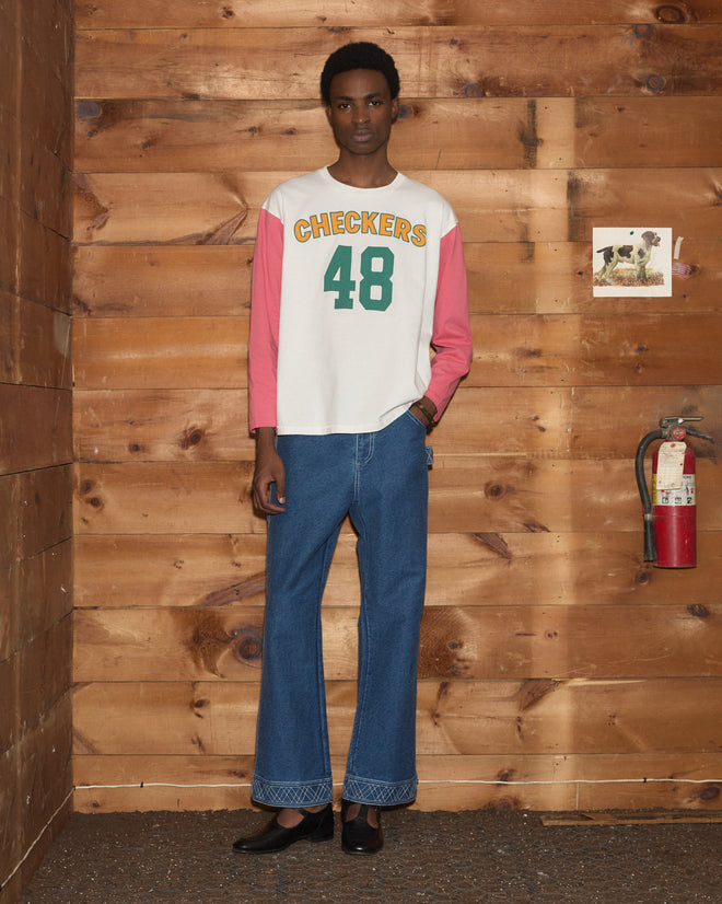 Wearing the Checkers Baseball Tee with flared jeans, a person stands by a wood wall adorned with a fire extinguisher and a dog photo, highlighting the casual style of this 100% cotton garment.