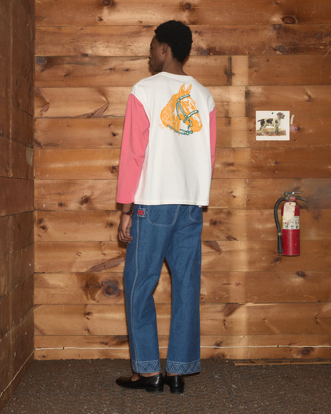 A person stands facing a wooden wall, dressed in jeans and the Checkers Baseball Tee—a 100% cotton shirt featuring a horse illustration on the back.