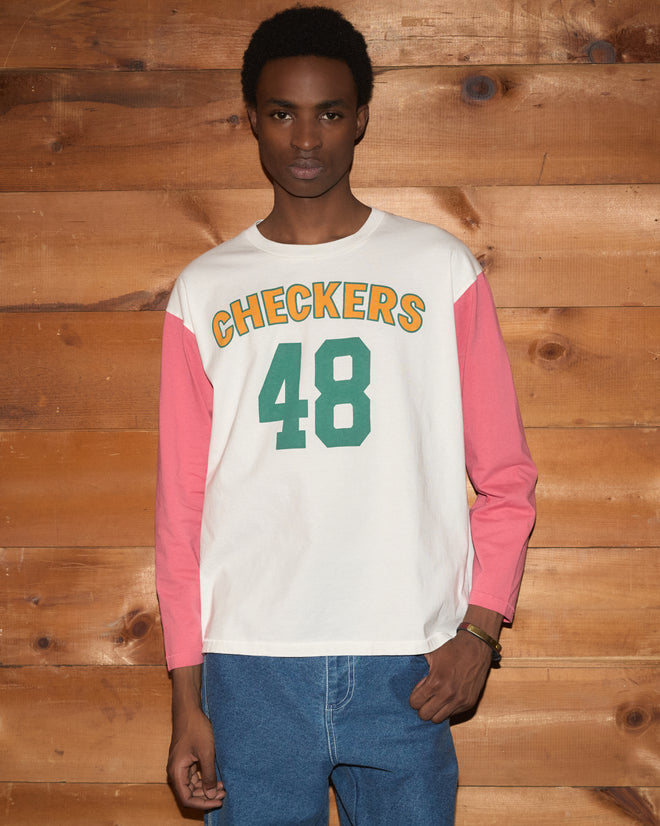 A person wearing the Checkers Baseball Tee—a 100% cotton top with pink sleeves and “CHECKERS 48” text—paired with blue jeans, stands by a wooden wall. This comfortable and stylish garment is perfect for any casual outing.