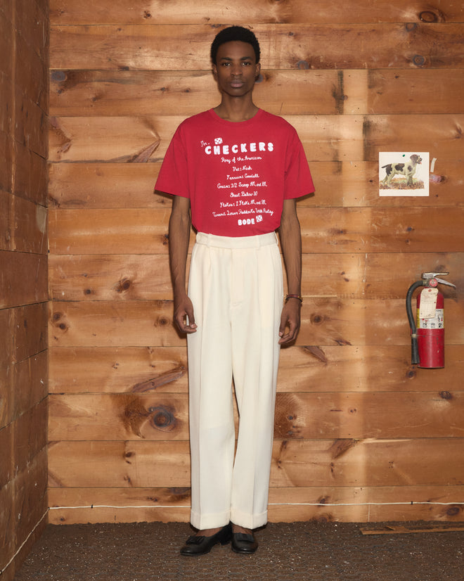 A person stands indoors by a wooden wall, wearing the red Feed Tee—crafted from 100% cotton—in a boxy fit, paired with cream pants and black shoes.