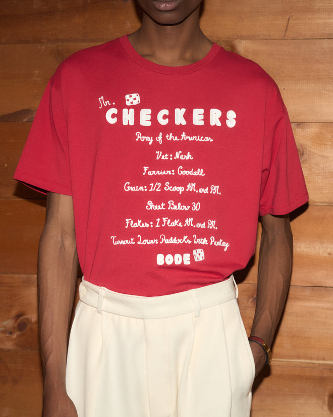 A person wearing the Feed Tee—a boxy, 100% cotton shirt in vibrant red featuring “CHECKERS” and playful white text—stands in front of a wooden wall. This tee combines comfort and style, making it perfect for any casual day.