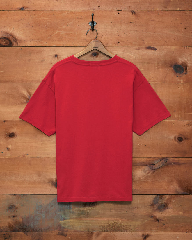 The Feed Tee, crafted from 100% cotton in a plain red boxy silhouette, hangs on a wooden hanger against a rustic wooden wall.